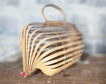 May include: A handcrafted, light brown bamboo basket with a curved handle. The basket is constructed from thin, parallel strips of bamboo, creating an open, airy design. A small, red square is attached to the bottom. The basket is likely used for carrying items.