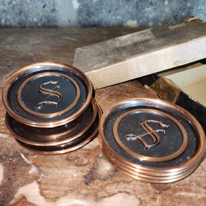 May include: Set of six round coasters with a copper-colored rim and a black center. Each coaster features a copper-colored letter "S" in the center. The coasters are stacked and arranged on a wooden surface, with a vintage box in the background.