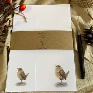 Peut inclure: Une pile de papier à lettres blanc ornée d'une illustration de petit oiseau brun au bas de chaque feuille. Une bande de papier brun entoure le centre avec le texte Pencil Art by MAGS et Writing Paper. Un stylo plume noir est posé à côté.
