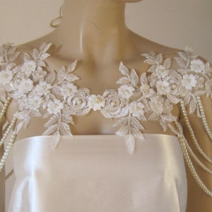 May include: A white lace bridal shoulder piece with pearl beading. The lace features delicate floral designs and the pearl strands hang down the shoulders.