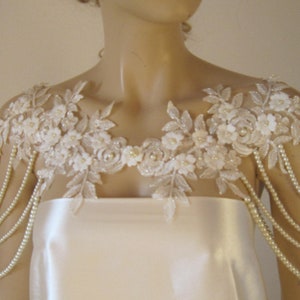 May include: A white lace bridal shoulder piece with pearl strands. The lace features delicate floral designs and is accented with small white pearls.