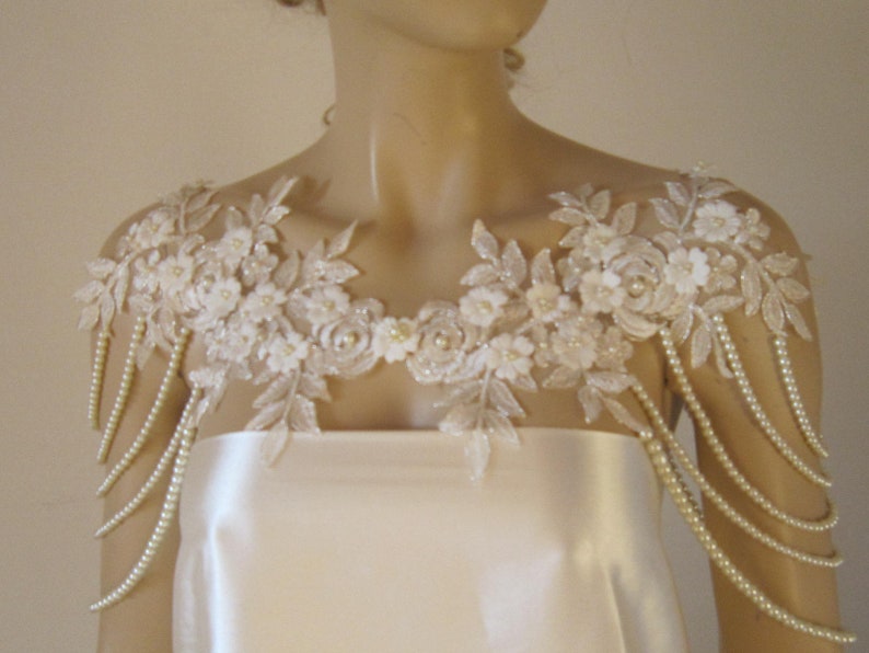May include: A white lace bridal shoulder piece with pearl strands. The lace features delicate floral designs and is accented with small white pearls.