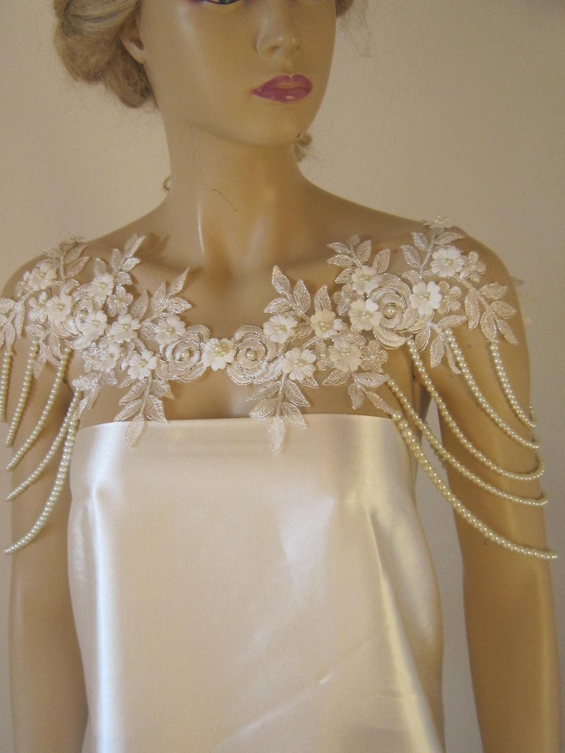 May include: A white lace bridal shoulder piece with pearl beading. The piece is designed to be worn over a strapless dress.