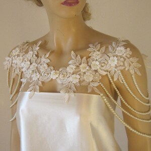 May include: A white lace bridal shoulder piece with pearl strands. The lace features delicate floral designs and is accented with small pearl beads.
