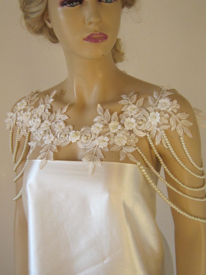 May include: A white lace bridal shoulder piece with pearl strands. The lace features delicate floral designs and is accented with small pearl beads.
