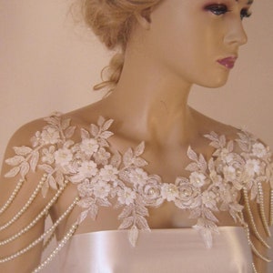 May include: A white lace bridal shoulder piece with pearl strands. The lace features delicate floral designs and is accented with small white pearls.