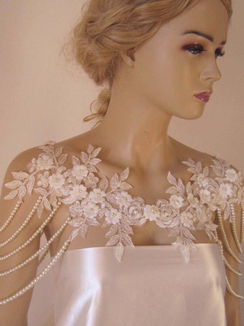May include: A white lace bridal shoulder piece with pearl strands. The lace features delicate floral designs and is accented with small white pearls.