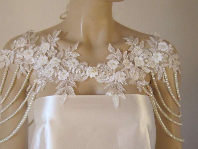 May include: A white lace bridal shoulder piece with pearl beading. The lace features delicate floral designs and the pearl strands hang down the shoulders.