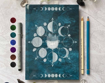 Puede incluir: Un diario azul turquesa con un diseño de fases lunares blancas y un caldero lleno de cristales. El diario tiene una superficie texturizada y un borde blanco alrededor de los bordes.