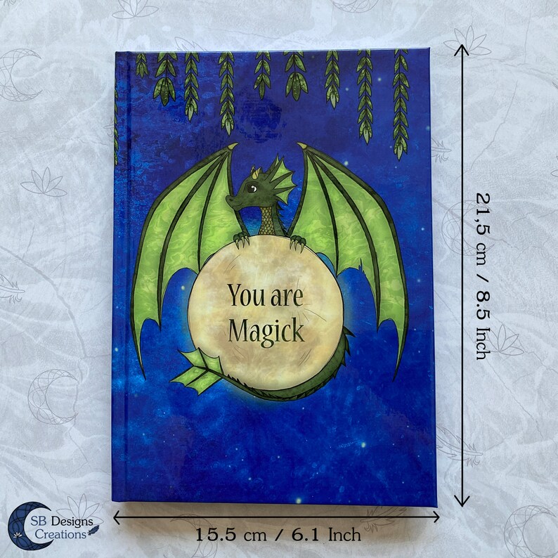 May include: A green dragon with gold accents is holding a moon-shaped sign that says "You are Magick". The dragon is on a blue background with stars and green leaves hanging from the top. The journal is 8.5 inches tall and 6.1 inches wide.