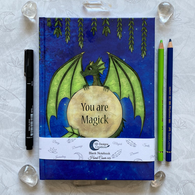 May include: A blue and green notebook with a dragon design and the text "You are Magick". The notebook is hardbound and has a white band around it with the text "SB Designs Creations, Blank Notebook, Hard Cover A5".