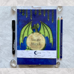 May include: A blue notebook with a green dragon illustration and the text "You are Magick". The notebook is decorated with stars and moon imagery. The notebook is labeled "Blank Notebook Hard Cover A5".