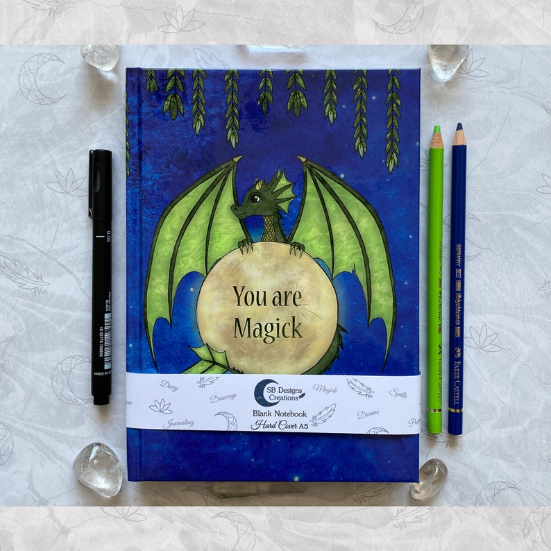 May include: A blue notebook with a green dragon illustration and the text "You are Magick". The notebook is decorated with stars and moon imagery. The notebook is labeled "Blank Notebook Hard Cover A5".