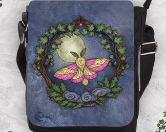 May include: A black messenger bag featuring a flap with a graphic illustration of a pink and yellow moth centered before a full moon. The moth is framed by a circular wreath of green leaves and brown branches, with gray rocks below and the text Sylluna.