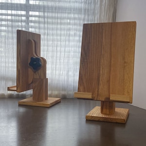 May include: Two wooden book stands. One stand has an adjustable angle with a black knob. The other stand is fixed. Both stands are made of light brown wood and have a rectangular base. The stands are on a dark brown surface.