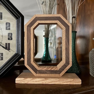 May include: A wooden octagonal picture frame with a dark brown inner border and a light brown outer border, mounted on a matching wooden base. The frame is displayed on a dark surface, with a clock and decorative vases in the background.