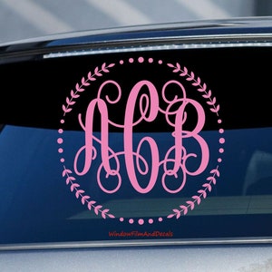 May include: Pink monogram decal with the letters ACB in a decorative script font. The monogram is surrounded by a wreath of leaves and a dotted circle. The decal is on a black car window.
