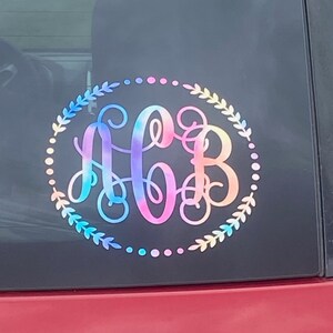 May include: A colorful monogram decal with the letters "AGB" in a rainbow tie-dye design. The monogram is surrounded by a wreath of leaves and a dotted circle.