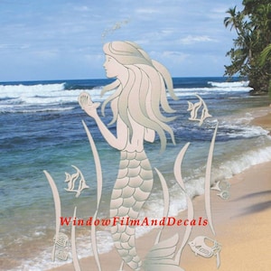 May include: A silver-colored mermaid with long flowing hair holding a seashell, surrounded by seaweed and fish. The mermaid is standing on a sandy beach with ocean waves in the background. The text "WindowFilmAndDecals" is visible at the bottom of the image.