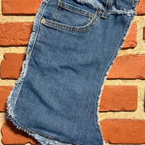 May include: A denim stocking with a frayed edge design, hanging against a red brick wall. The stocking is made from blue jean material, featuring a pocket and belt loops. A unique holiday decoration.