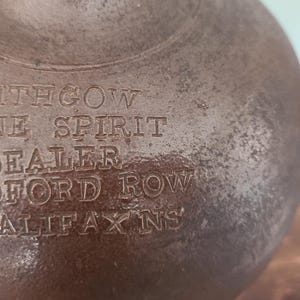 May include: Close-up of a brown, antique, spherical container with engraved text. The text reads "J LITHGOW WINE SPIRIT DEALER BEDFORD ROW HALIFAX NS". The container has a textured surface and appears aged.