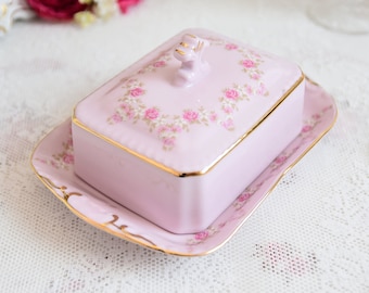 May include: A pink porcelain butter dish with a floral design and a lid featuring a gold trim. The dish is sitting on a white lace tablecloth.