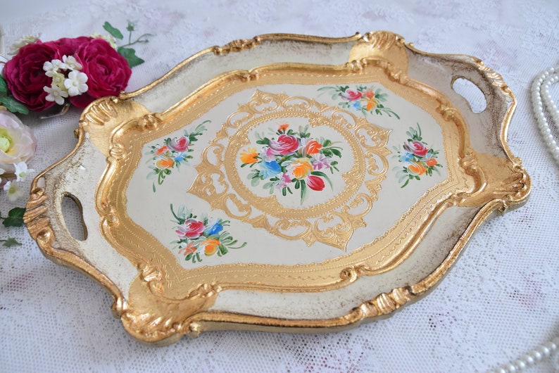 May include: A white and gold serving tray with ornate details and painted floral designs. The tray has two handles and a scalloped edge.