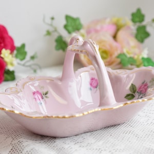 May include: A pink porcelain dish with a handle in the shape of a swan. The dish is decorated with gold trim and floral designs.
