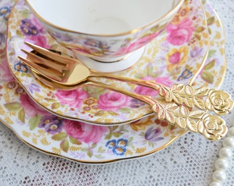 May include: A pair of gold-tone teaspoons with rose-shaped handles. The spoons are resting on a floral patterned teacup and saucer.