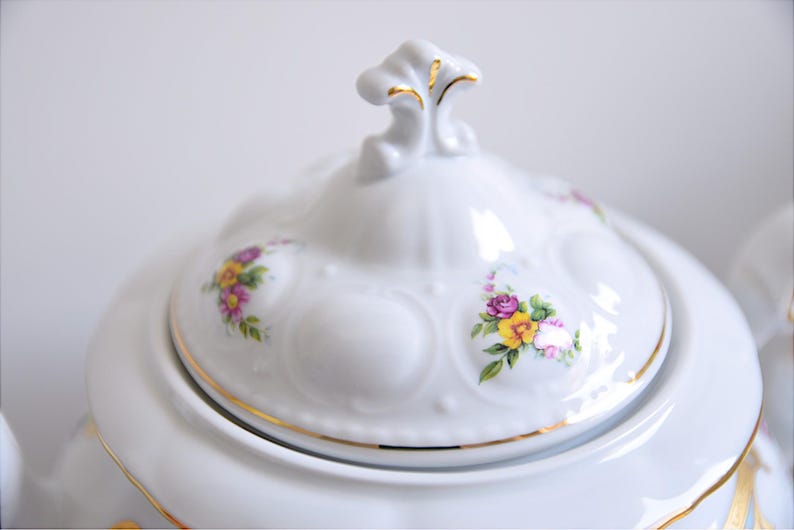 May include: White porcelain teapot with a floral design. The lid has a gold trim and a decorative knob. The teapot has a floral pattern with pink, yellow, and purple flowers.