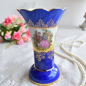 May include: A cobalt blue porcelain vase with a gold trim and a painted scene of a couple in a garden. The vase has a scalloped rim and a flared base.