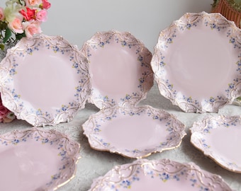 May include: Set of seven pink and gold decorative plates with a floral design. The plates have a scalloped edge and are decorated with blue and gold flowers.