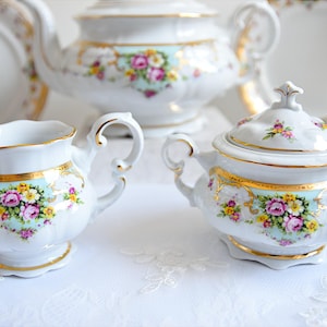 May include: A vintage floral tea set with a teapot, creamer, sugar bowl, and cups. The set is white with gold trim and features a delicate floral pattern in shades of pink, yellow, and blue.
