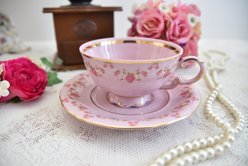 Puede incluir: Una taza de t&eacute; y un platillo rosas con un patr&oacute;n floral y ribete dorado. La taza de t&eacute; y el platillo est&aacute;n sobre un mantel de encaje blanco con una cadena de perlas.