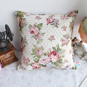 May include: A square throw pillow with a floral pattern featuring pink, red, and white roses on a cream background with small white polka dots. The pillow has a white border.