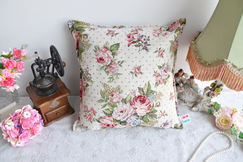 May include: A square throw pillow with a floral pattern featuring pink, red, and white roses on a cream background with small white polka dots. The pillow has a white border.