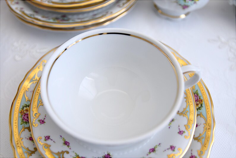 May include: A white teacup with a gold rim and a floral pattern on the saucer. The teacup is empty and the saucer has a light blue background.