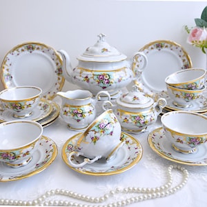 May include: A vintage porcelain tea set with floral and gold accents. The set includes a teapot, creamer, sugar bowl, and four teacups and saucers.