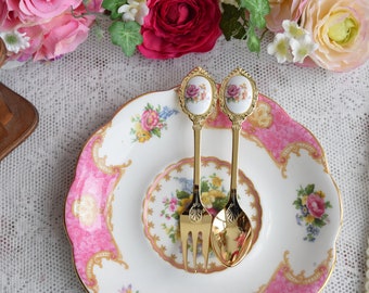 May include: Two gold-tone teaspoons with floral designs on the handles. The spoons are on a pink and white floral patterned plate.