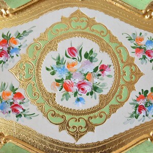 K&ouml;nnte beinhalten: Ein dekoratives Tablett mit einem gr&uuml;n-goldenen Design. Das Tablett zeigt ein florales Motiv mit rosa, wei&szlig;en und gelben Blumen. Das Tablett ist oval geformt und hat einen erh&ouml;hten Rand.