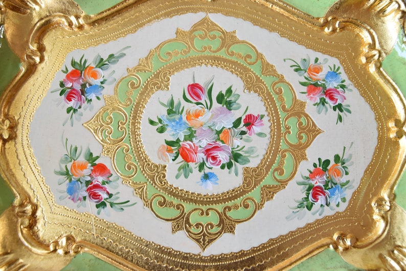 K&ouml;nnte beinhalten: Ein dekoratives Tablett mit einem gr&uuml;n-goldenen Design. Das Tablett zeigt ein florales Motiv mit rosa, wei&szlig;en und gelben Blumen. Das Tablett ist oval geformt und hat einen erh&ouml;hten Rand.