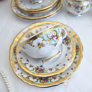May include: A vintage teacup and saucer set with a floral pattern and gold trim. The cup and saucer are white with a light blue background and feature pink, yellow, and green flowers. The gold trim is intricate and adds a touch of elegance to the set.