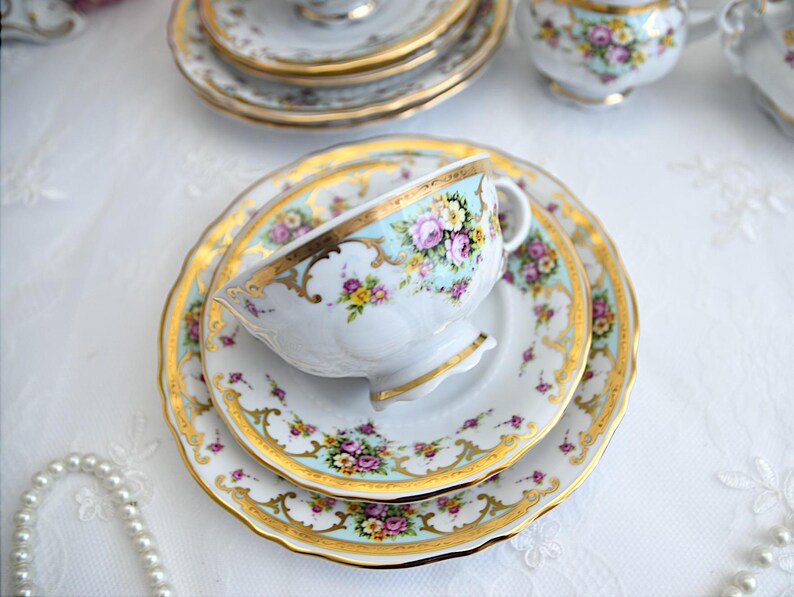 May include: A vintage teacup and saucer set with a floral pattern and gold trim. The cup and saucer are white with a light blue background and feature pink, yellow, and green flowers. The gold trim is intricate and adds a touch of elegance to the set.