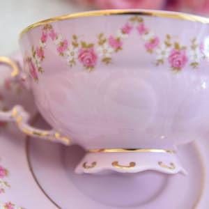 Puede incluir: Un delicado juego de taza y platillo de t&eacute; de color lavanda con un patr&oacute;n floral de rosas rosas y flores blancas. El juego tiene un borde dorado.
