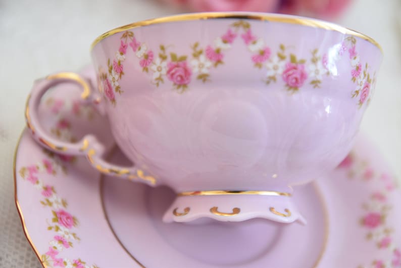 Puede incluir: Un delicado juego de taza y platillo de t&eacute; de color lavanda con un patr&oacute;n floral de rosas rosas y flores blancas. El juego tiene un borde dorado.