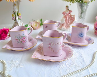 May include: A set of four pink heart-shaped teacups and saucers with floral designs. The cups and saucers are decorated with delicate pink and blue flowers.