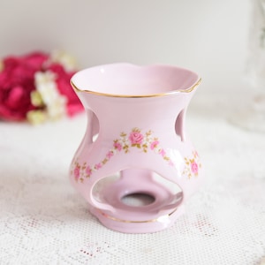 Könnte beinhalten: Ein rosa Keramikkamin mit floralem Design und goldener Verzierung. Der Kamin hat ein ausgeschnittenes Design an den Seiten und eine runde Öffnung in der Mitte.