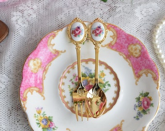 May include: Two gold-tone teaspoons with floral designs on the handles, resting on a pink and white floral patterned saucer.