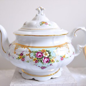May include: A white porcelain teapot with a floral design and gold accents. The teapot has a lid with a knob on top. The teapot is sitting on a white surface.