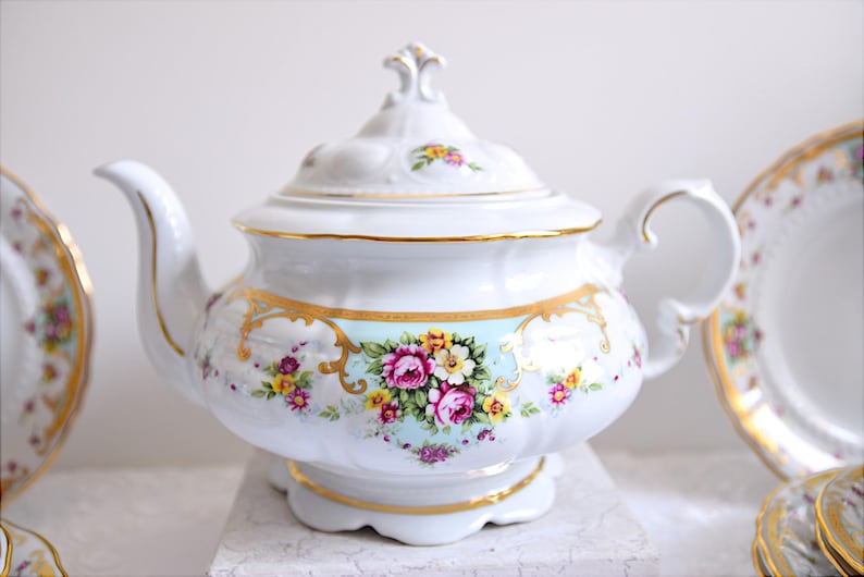May include: A white porcelain teapot with a floral design and gold accents. The teapot has a lid with a knob on top. The teapot is sitting on a white surface.